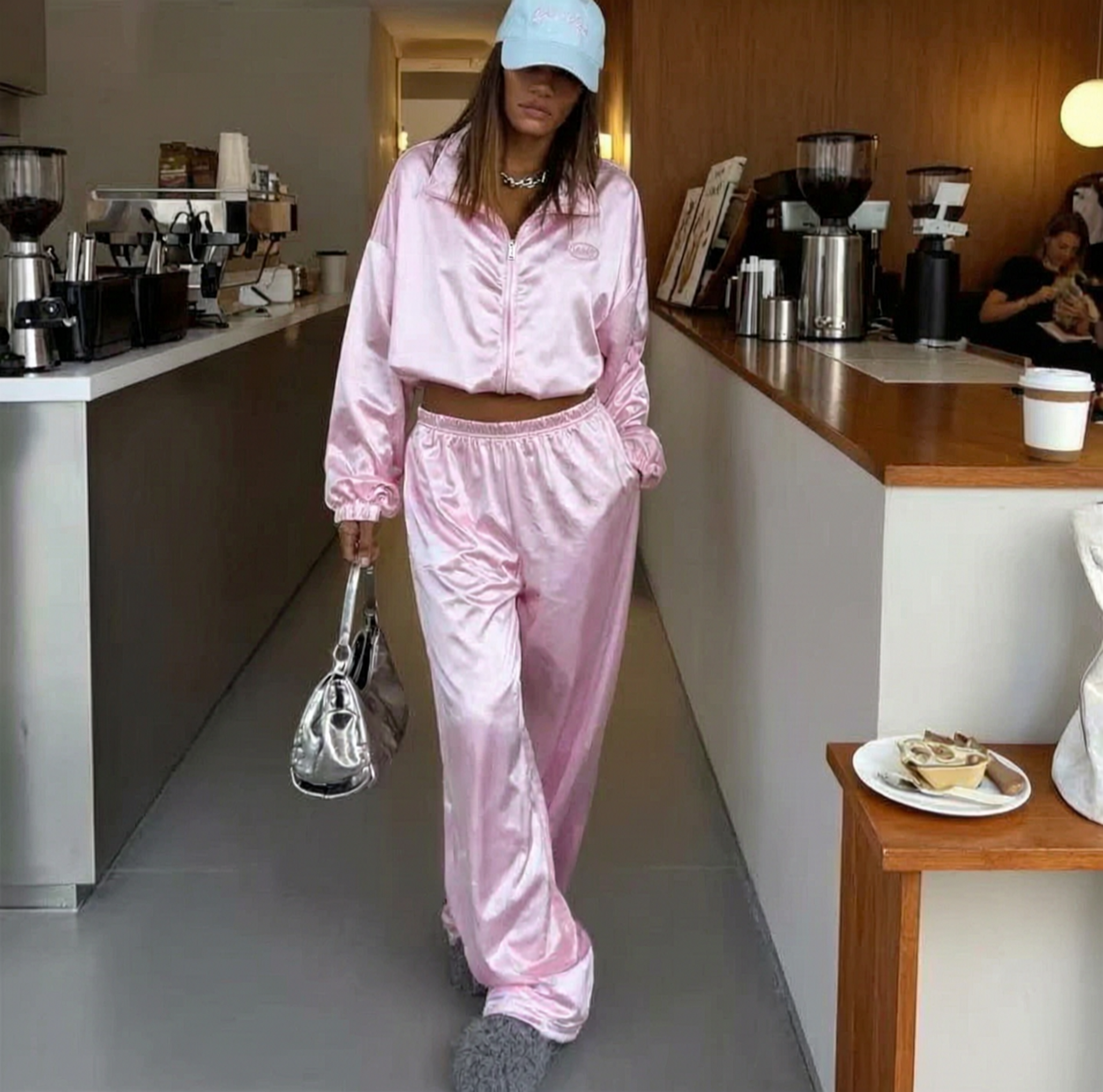 Model walking in the pink satin Soft Serve Set - long-sleeve top and wide-leg trousers - with a blue cap and silver handbag